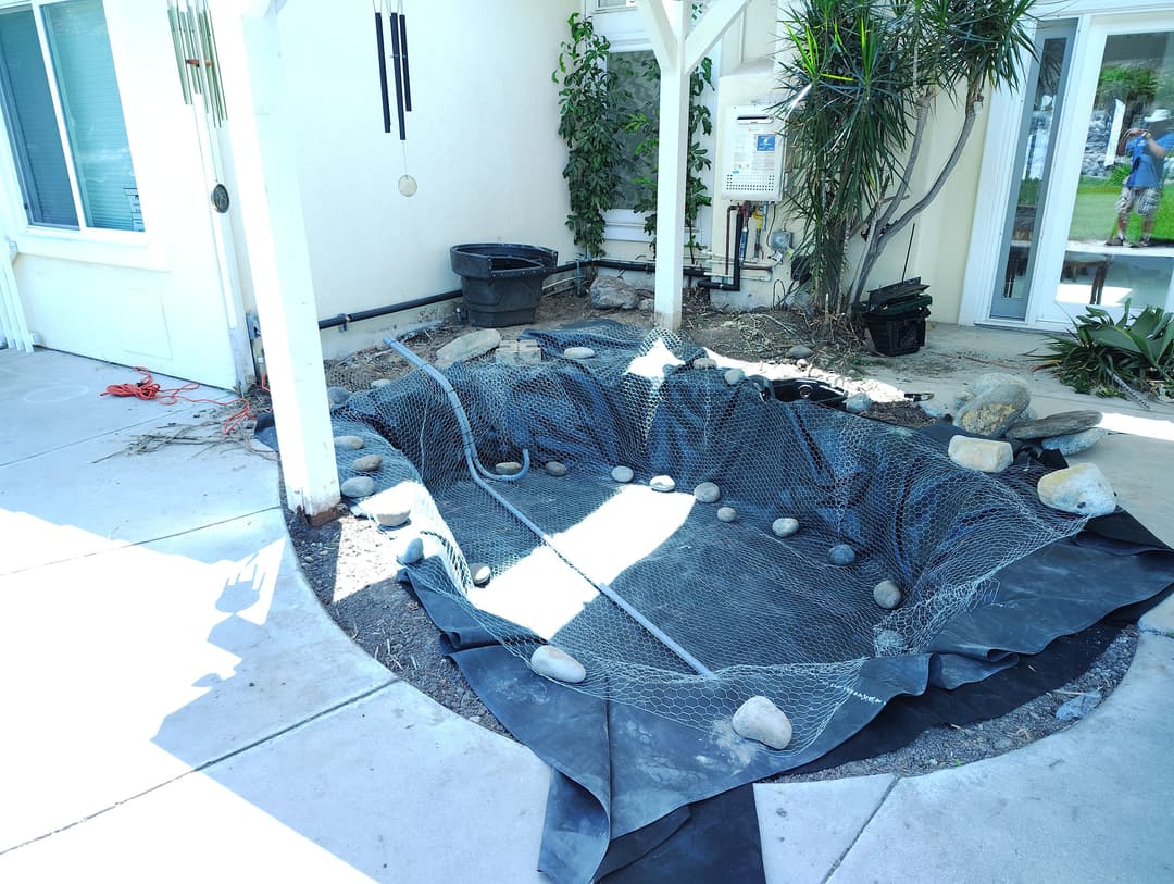 Pond construction site with protective mesh, landscaping stones, and garden equipment visible.