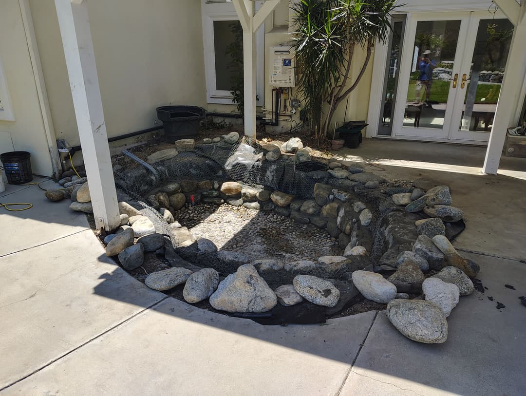 Outdoor pond area under construction with rocks and mesh around a gravel base.