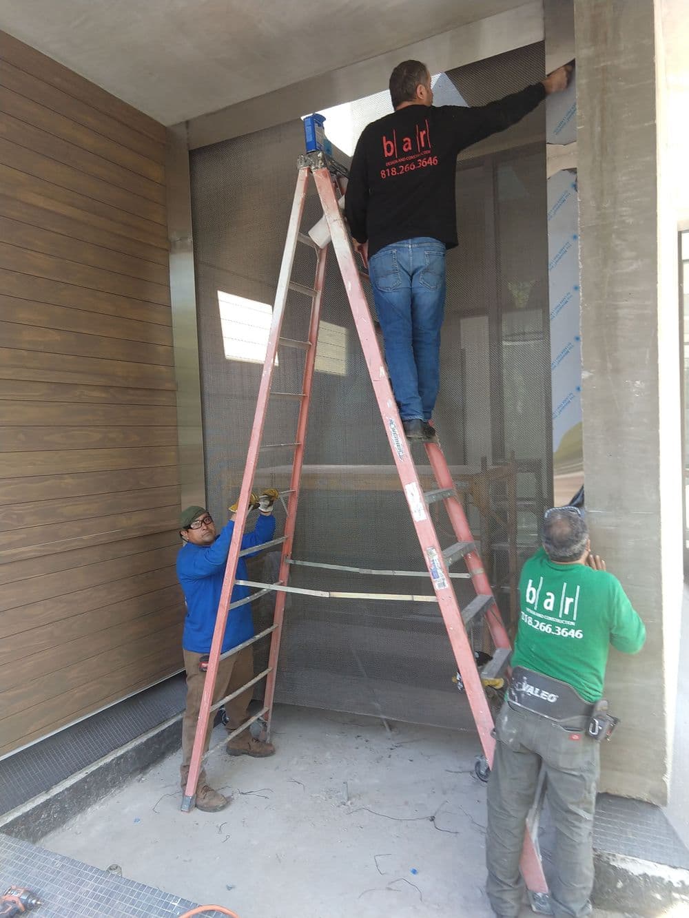 Workers installing a metal mesh screen on a building entrance using a ladder.