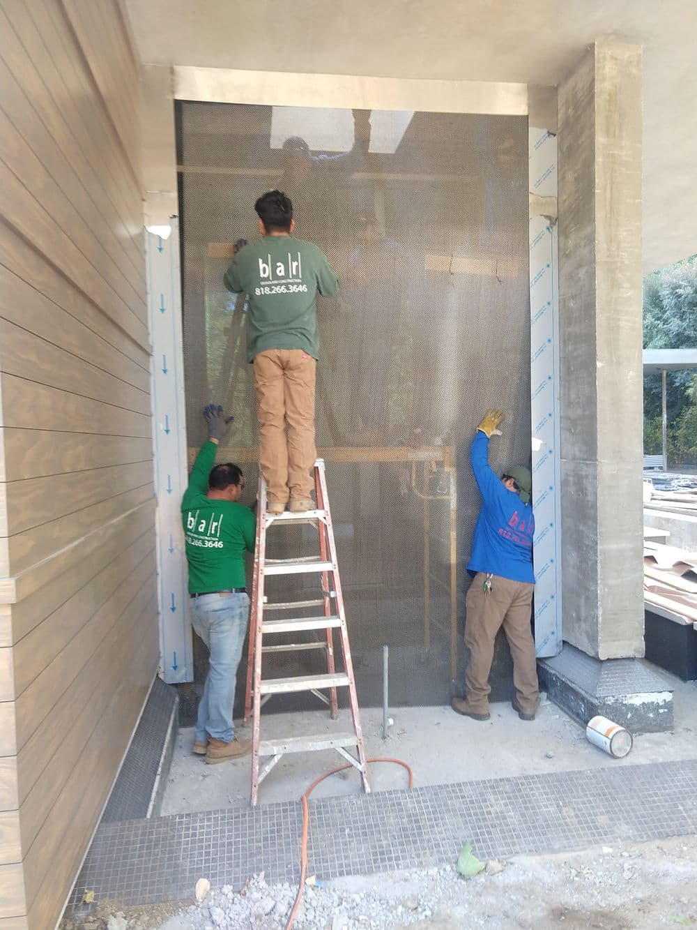 Workers installing mesh screen on a building exterior with ladder and construction materials nearby.