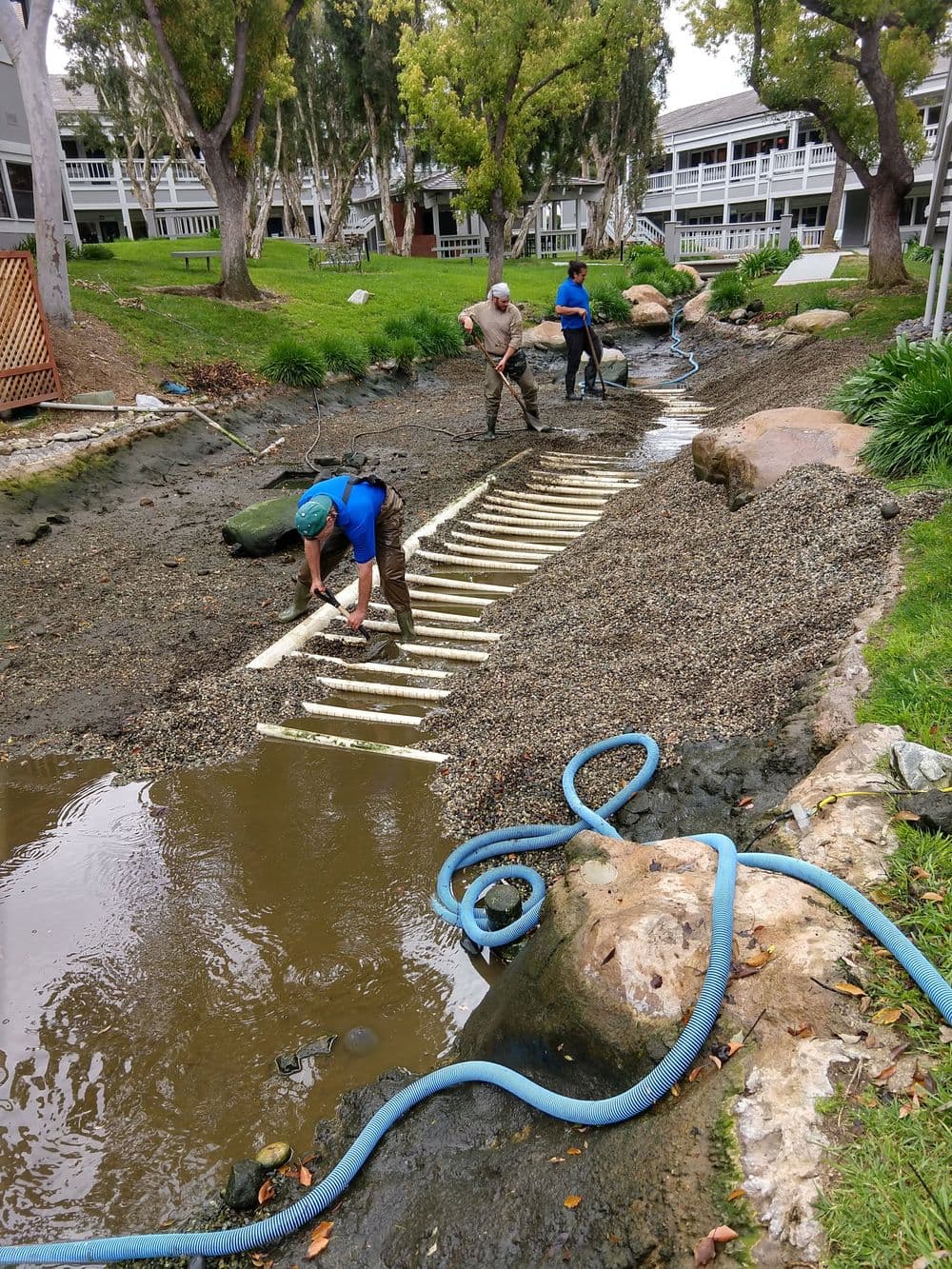 Workers constructing a wooden pathway along a waterway in a landscaped area.
