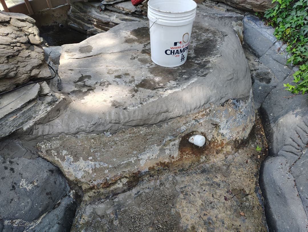 Bucket on rocky surface with deteriorating concrete and drainage pipe in a garden setting.