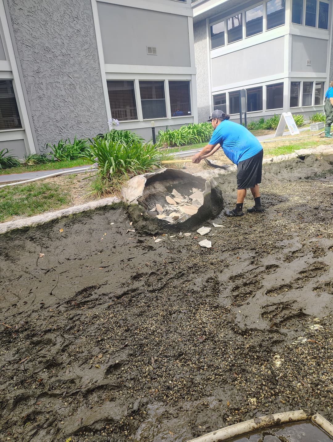 Man removing debris from a muddy area near a building, landscaping work in progress.