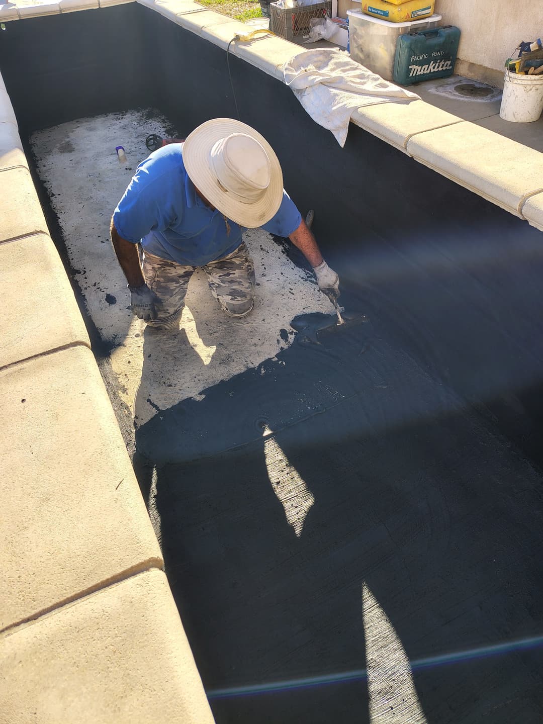 Worker applying sealant to an empty swimming pool, wearing a straw hat and blue shirt.