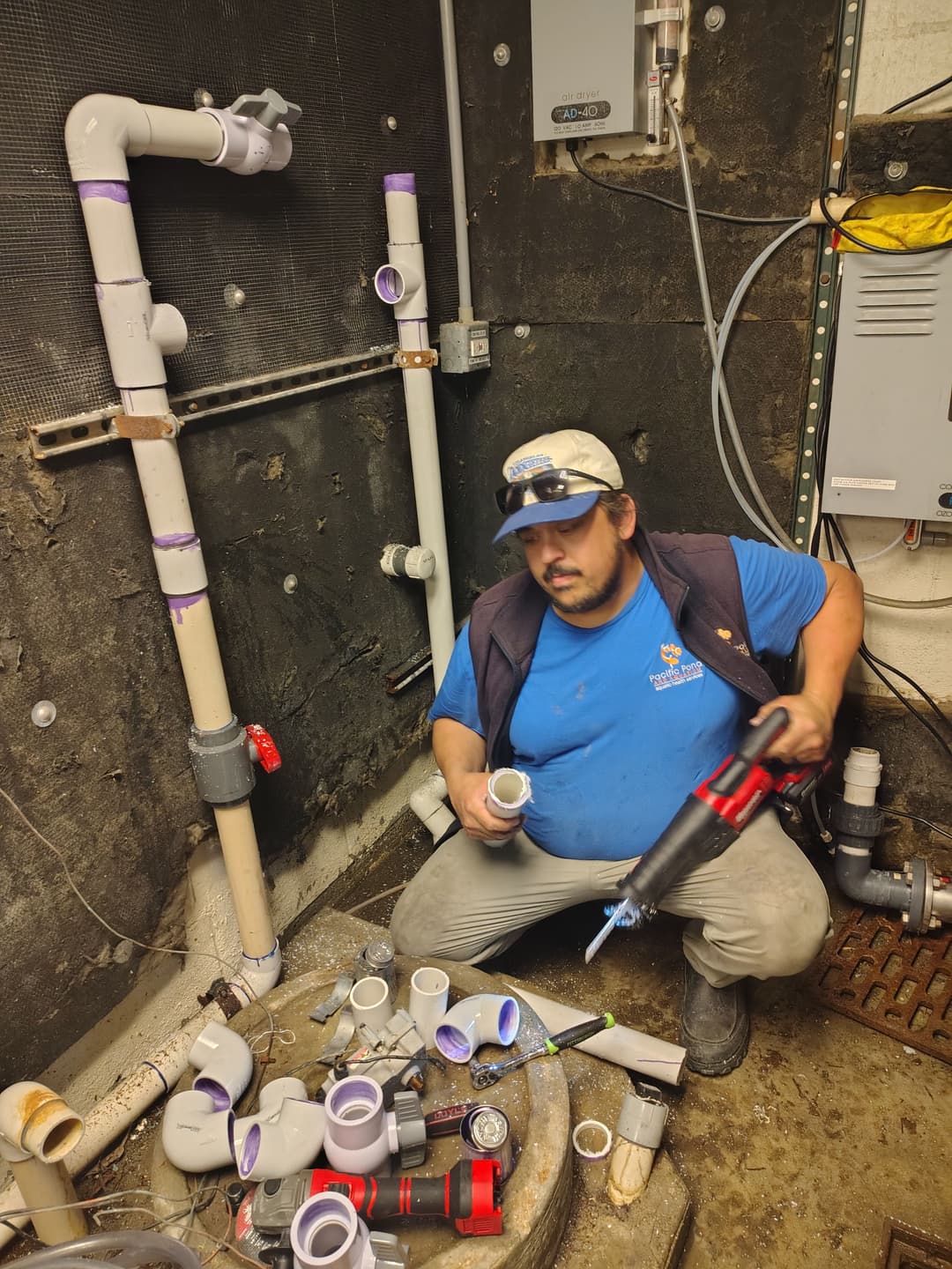 Plumber working on PVC pipe installation in a basement, surrounded by various piping materials.
