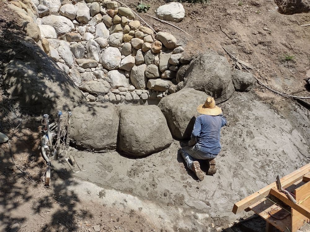 Person kneeling while applying concrete to stone wall in outdoor landscape project.