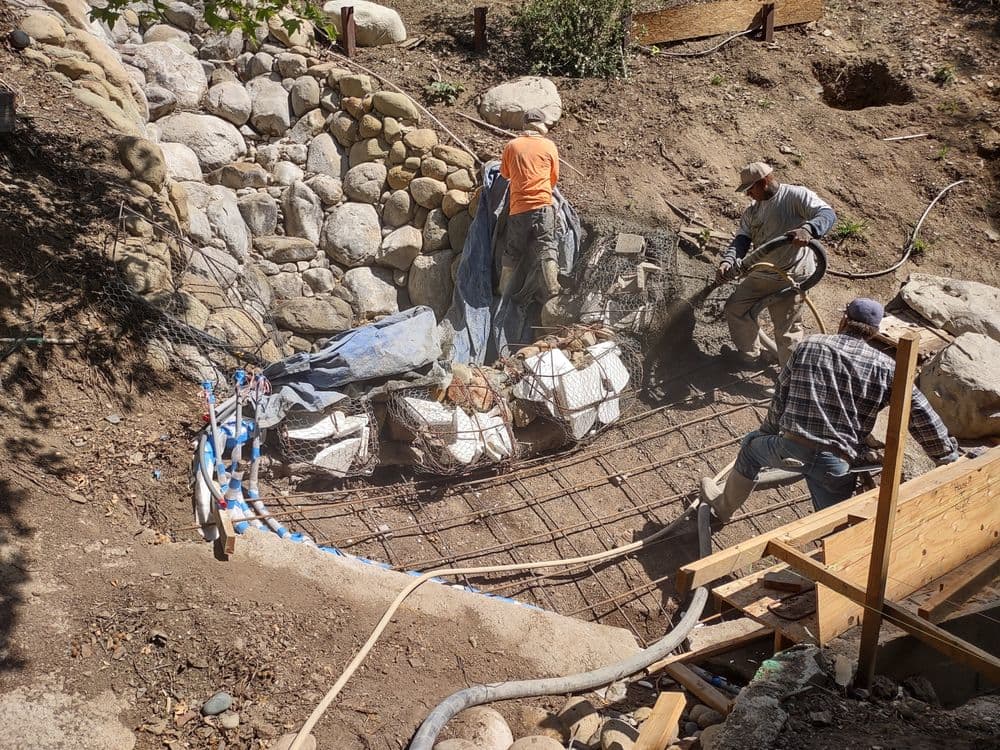 Construction workers reinforcing a stone wall with rebar and mesh in a rocky landscape.