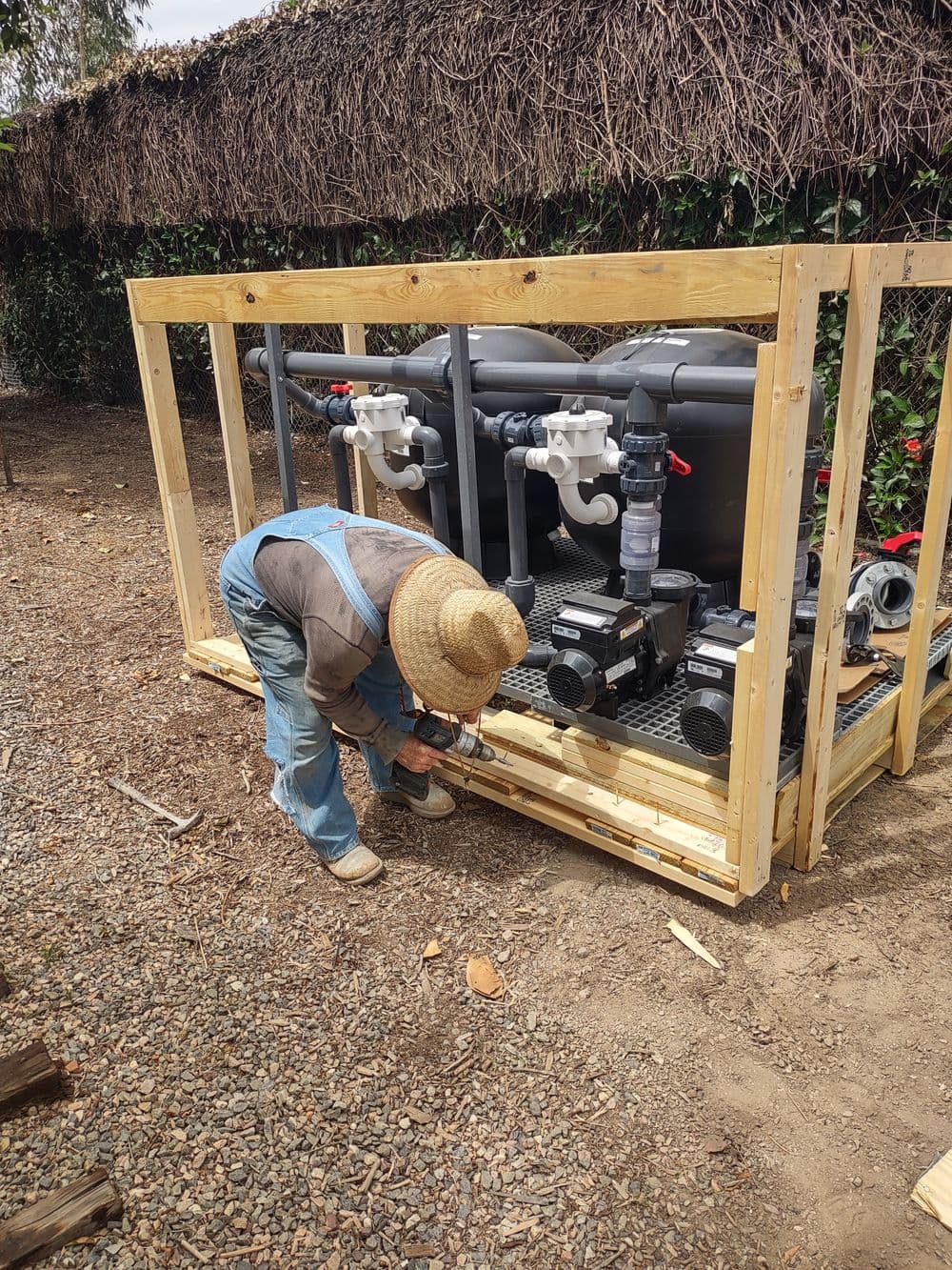 Worker installing pool equipment in wooden frame outdoors with tools and machinery nearby.