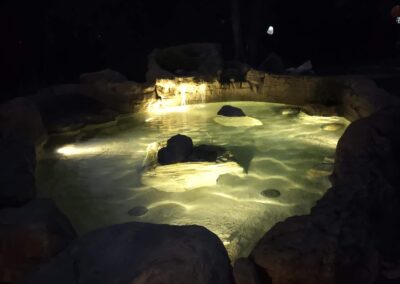 Illuminated outdoor hot tub with natural stone edges surrounded by trees at night.