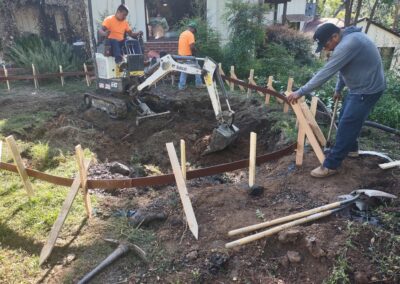Workers using heavy machinery to create a foundation in a garden area with wooden stakes.