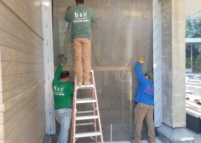 Workers installing mesh screen on a building exterior with ladder and construction materials nearby.