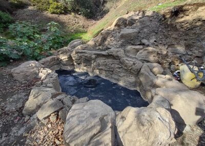 Natural stone hot spring pool surrounded by greenery and rocky terrain.