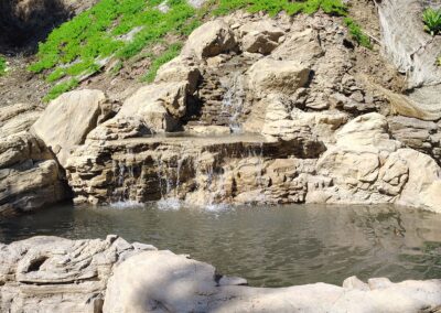 Natural rock waterfall with tranquil pool surrounded by lush green vegetation in a serene setting.
