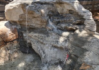 Artificial rock waterfall with water flowing, surrounded by a fenced area in a park setting.