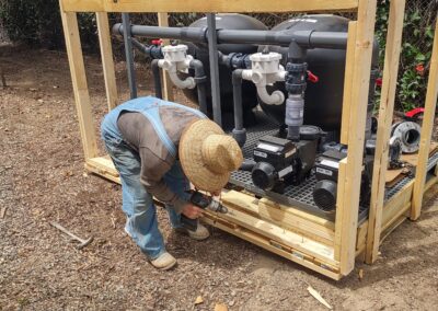 Worker assembling pool equipment in a wooden frame outdoors with tools and machinery visible.
